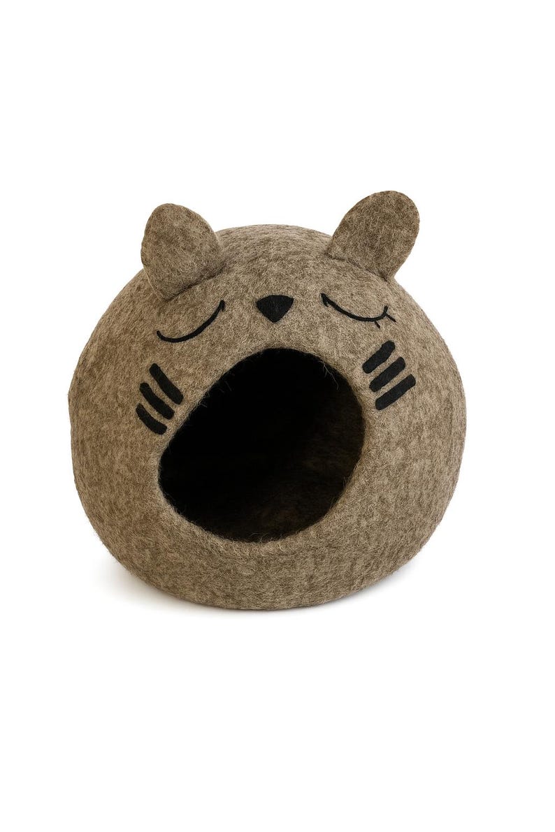 May include: A cat-shaped pet bed in a neutral brown color. The bed is made of felt and features a round shape with a large opening. Black details include closed eyes, a nose, and whiskers.