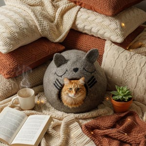 May include: A cozy scene featuring a gray cat bed shaped like a cat's head, with an orange cat inside. Surrounding the bed are cream and rust-colored pillows and blankets. A cup of coffee and a book are also present.