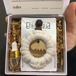 Könnte beinhalten: Eine weiße Schachtel mit einem Makramee-Anhänger mit Holzmitte und einer kleinen Glasflasche mit goldenem Verschluss. Der Anhänger hat ein kreisförmiges Design mit einem schwarzen und braunen Stein oben drauf.
