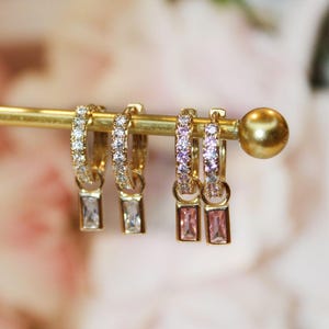 May include: Gold-toned hoop earrings with clear and pink rectangular pendants. Two pairs of earrings are displayed on a gold bar. One pair has clear stones, the other has pink stones. The hoops are encrusted with small, clear stones.