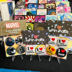 May include: A display of various pin packs and individual pins. The packs include "Marvel Bucky Barnes 4-Pack" and "2-Pin Pack" with star and heart designs. Individual pins feature text like "I Lewis Pullman" and "I Bob Reynolds."