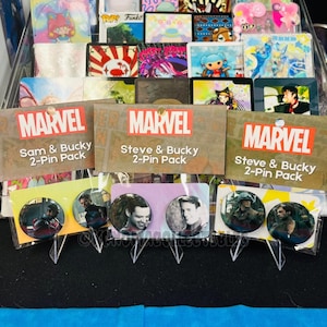 May include: Marvel 2-pin packs featuring Sam & Bucky and Steve & Bucky. Each pack includes two round pins with character images. The Marvel logo is displayed on the packaging. The background includes various collectible cards.