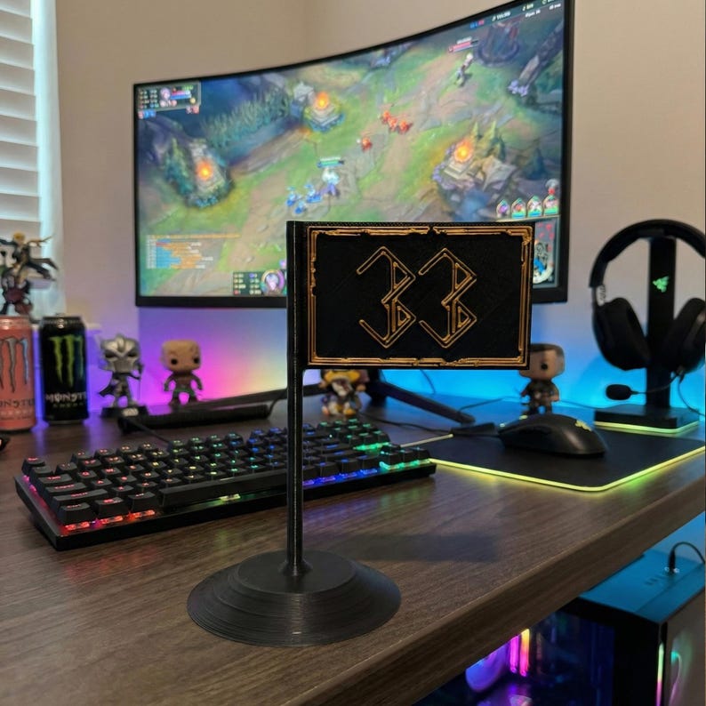 May include: A black desk flag with the number "33" in gold, set against a gaming setup. The flag is rectangular with a gold border and a black background. The base is round. A curved monitor and keyboard are in the background.