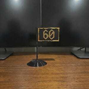 May include: A black and gold flag with the number 60 in a stylized font. The flag is mounted on a black stand. The flag is likely a decorative item or a marker.