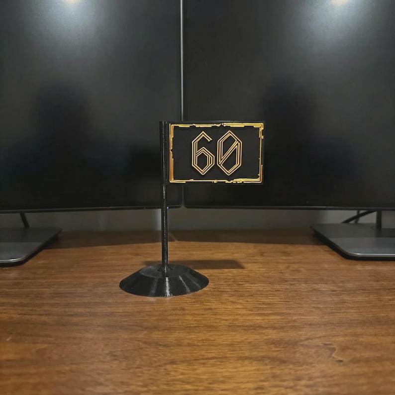 May include: A black and gold flag with the number 60 in a stylized font. The flag is mounted on a black stand. The flag is likely a decorative item or a marker.
