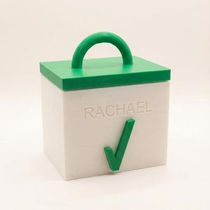 Puede incluir: Una caja cuadrada blanca con tapa y asa verdes. El nombre "RACHAEL" está grabado en la parte delantera, junto con una marca de verificación verde. El asa tiene forma arqueada.