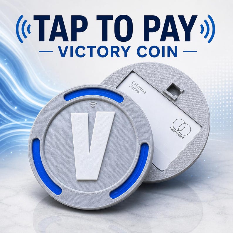May include: A circular gray Victory Coin with a large white letter V and blue accents, featuring a contactless payment symbol. The coin is positioned in front of a matching gray circular holder containing a white California Series Mastercard.