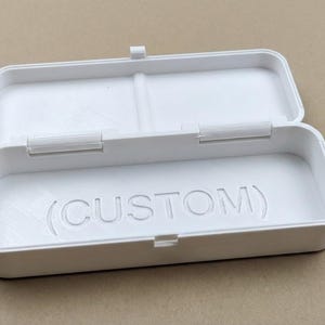 Puede incluir: Caja rectangular blanca abierta con tapa con bisagras. La palabra "(CUSTOM)" está grabada en el interior. La caja es de un material liso y mate. La tapa está abierta, revelando el interior.