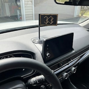 May include: A black and gold car flag with the number "33" displayed on the flag. The flag is mounted on a silver pole and base, placed on a car dashboard. The car interior is visible, including the steering wheel and infotainment screen.