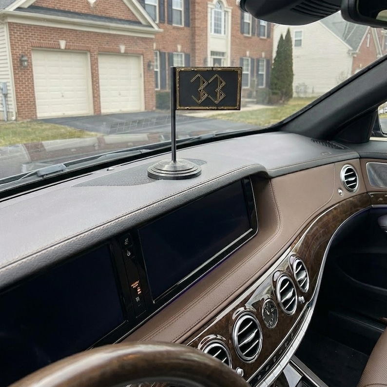 May include: A small black flag with the number "33" in gold lettering, mounted on a silver stand. The flag is displayed on the dashboard of a car with a brown interior. The car is parked in front of a house.