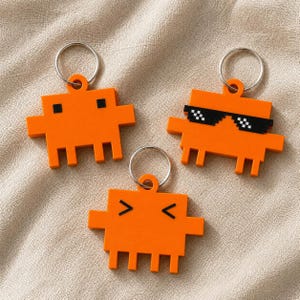 May include: Three orange pixelated character keychains with silver metal rings. One has black square eyes, one wears black pixelated sunglasses, and one has black chevron-shaped eyes. Each figure has a rectangular body with side arms and four legs.