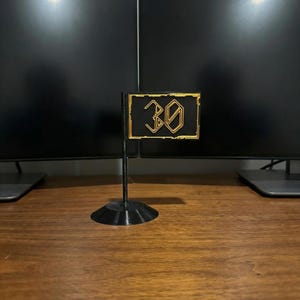 May include: A black and gold flag-shaped desk accessory with the number "30" in a stylized font. The flag is mounted on a black stand. The background includes two black computer monitors and a wooden desk surface.