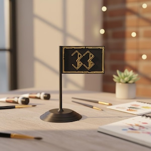 May include: A black table flag with the number "33" in gold, set on a round base. The flag is on a table with paintbrushes, a small succulent plant, and art supplies. The background includes a brick wall and a window.