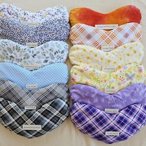 May include: A collection of ten, heart-shaped, fabric eye pillows in various patterns and colors. Patterns include floral, plaid, and abstract designs. Each pillow has a label with a descriptive name, such as "Wildflowers" or "Purple Plaid."