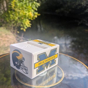 Könnte beinhalten: Eine weiße, rechteckige Box mit dem Logo der University of Michigan und einer Footballhelm-Grafik. Das Logo ist in Gelb und Marineblau gehalten, mit dem Aufdruck "MICHIGAN". Die Box steht auf einer Glasoberfläche.