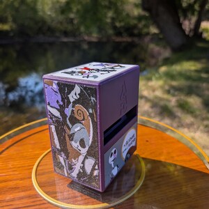 May include: A purple box featuring characters and scenes from "The Nightmare Before Christmas." The box has a lid with a design and the sides display images of Jack Skellington and other characters. The box is sitting on a wooden surface.