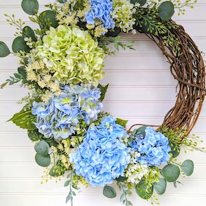 May include: A decorative wreath featuring a circular grapevine base adorned with vibrant blue and green hydrangeas, white flowers, and green foliage. The wreath is set against a white background.