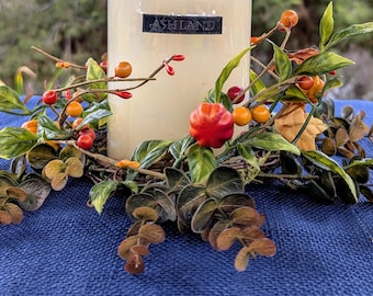 May include: A cream-colored pillar candle with the word "ASHLAND" on the front, surrounded by a decorative wreath. The wreath features green leaves, orange berries, and red accents, all set against a blue surface.