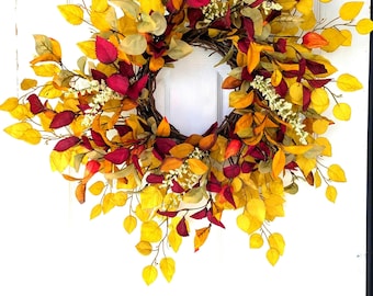 May include: A circular wreath constructed from a woven twig base, densely decorated with artificial autumn foliage. The arrangement features a vibrant mix of bright yellow, deep burgundy, and muted green leaves, accented by small clusters of white berries.