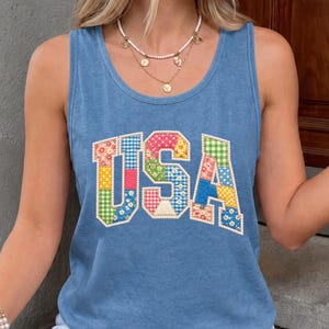 May include: A blue sleeveless tank top featuring the letters USA in a patchwork design. The letters are composed of various colorful fabric patterns including gingham, floral, and polka dots, outlined with white stitching on a solid blue background.