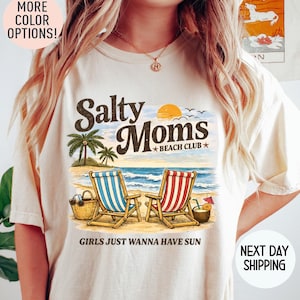 Op de afbeelding: Crèmekleurig T-shirt met een strandscène-afbeelding. Het ontwerp bevat de tekst "Salty Moms Beach Club" met palmbomen, strandstoelen en de zin "Girls Just Wanna Have Sun."