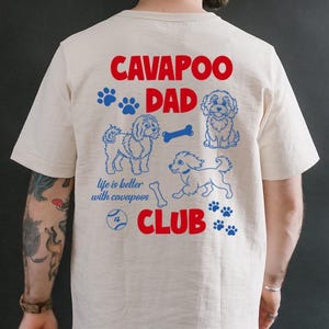 May include: Cream-colored t-shirt with a red and blue graphic that reads "CAVAPOO DAD CLUB." The design includes paw prints, dog illustrations, and the phrase "life is better with cavapoos."