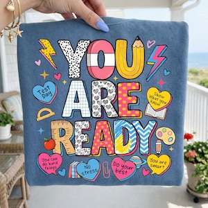 May include: A blue sweatshirt with the words "YOU ARE READY" in colorful, playful lettering. The design includes lightning bolts, hearts, and other school-themed graphics. The sweatshirt is held up by a person.