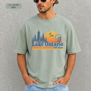 May include: A sage green short-sleeve t-shirt featuring a graphic print of a lighthouse, pine trees, a wave, and birds against a yellow and orange sunset. The text reads Lake Ontario EST 1656. The shirt has a crew neckline and a relaxed fit.