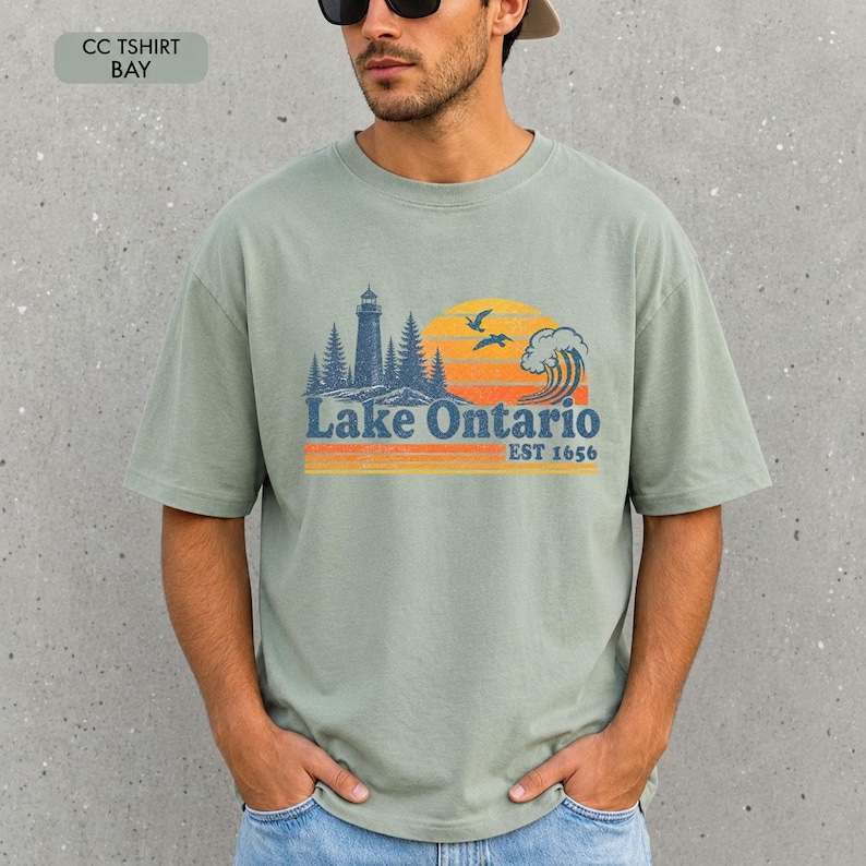 May include: A sage green short-sleeve t-shirt featuring a graphic print of a lighthouse, pine trees, a wave, and birds against a yellow and orange sunset. The text reads Lake Ontario EST 1656. The shirt has a crew neckline and a relaxed fit.