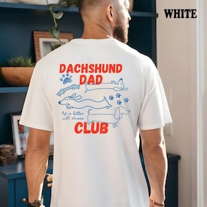 May include: White t-shirt featuring a dachshund-themed design. The graphic includes the words "DACHSHUND DAD CLUB" in red, accompanied by blue illustrations of dachshunds, paw prints, and a bone. The phrase "life is better with doxies" is also present.