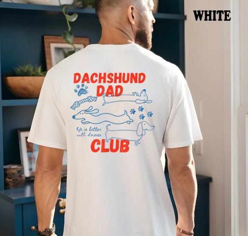 May include: White t-shirt featuring a dachshund-themed design. The graphic includes the words "DACHSHUND DAD CLUB" in red, accompanied by blue illustrations of dachshunds, paw prints, and a bone. The phrase "life is better with doxies" is also present.