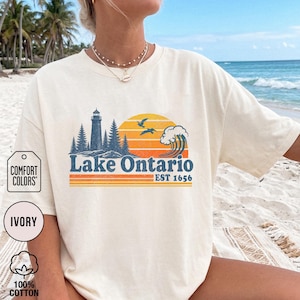 May include: An ivory t-shirt featuring a graphic of a lighthouse, pine trees, a sunset, birds, and a wave. The text reads Lake Ontario EST 1656. The design uses blue, orange, and yellow colors. The shirt has a crew neckline and short sleeves.