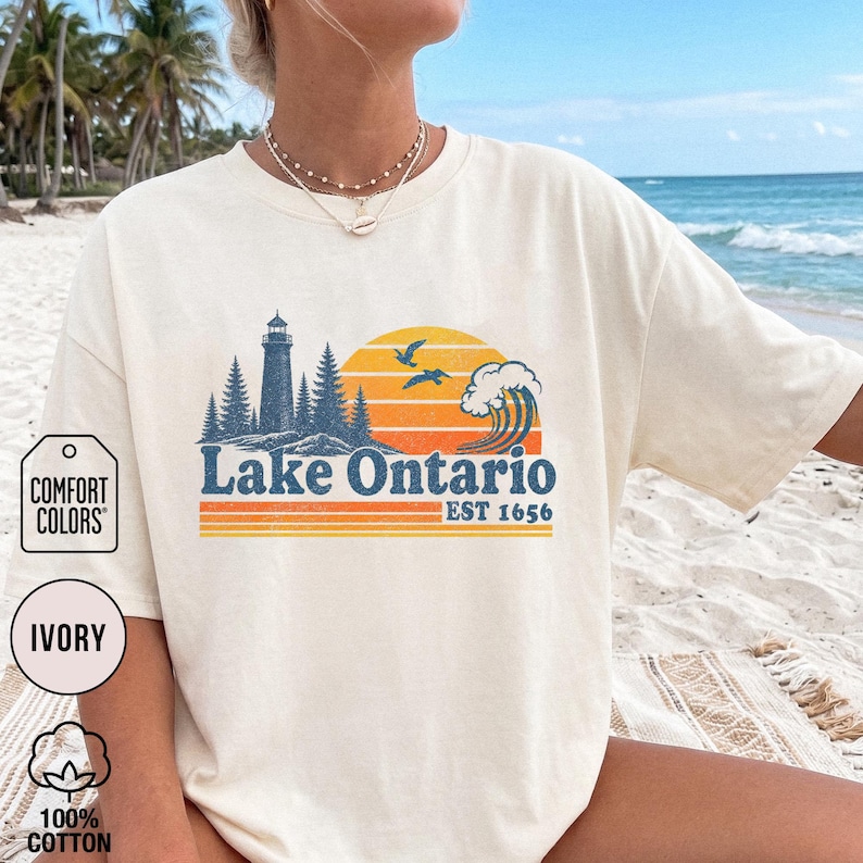 May include: An ivory t-shirt featuring a graphic of a lighthouse, pine trees, a sunset, birds, and a wave. The text reads Lake Ontario EST 1656. The design uses blue, orange, and yellow colors. The shirt has a crew neckline and short sleeves.