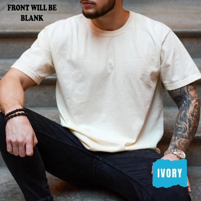 May include: Ivory-coloured short-sleeved t-shirt. The text "FRONT WILL BE BLANK" is at the top. The word "IVORY" is in a blue box at the bottom. The shirt is being worn by a person wearing black jeans and a black bracelet.