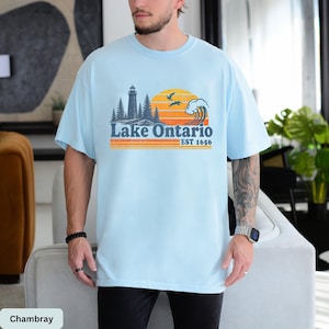 May include: A light blue t-shirt featuring a graphic print of a lighthouse, pine trees, a large sun, and a crashing wave. The text reads Lake Ontario EST 1656 in dark blue and orange, with horizontal orange and yellow stripes below the text.