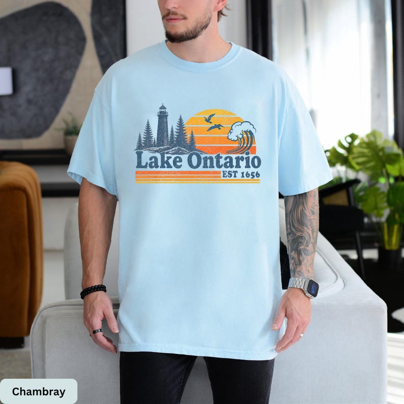 May include: A light blue t-shirt featuring a graphic print of a lighthouse, pine trees, a large sun, and a crashing wave. The text reads Lake Ontario EST 1656 in dark blue and orange, with horizontal orange and yellow stripes below the text.