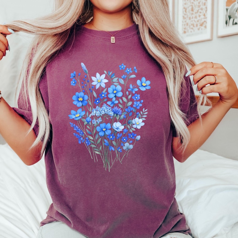 May include: A maroon t-shirt featuring a floral design. The graphic showcases a bouquet of blue flowers with green stems and leaves. The flowers vary in shades of blue, creating a visually appealing contrast against the shirt's background.