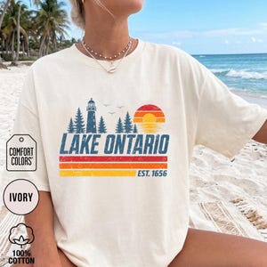 May include: An ivory short-sleeve t-shirt featuring a vintage-style graphic of a lighthouse, pine trees, and a setting sun. The text reads LAKE ONTARIO and EST. 1656, accompanied by horizontal red, orange, and yellow stripes with a distressed texture.