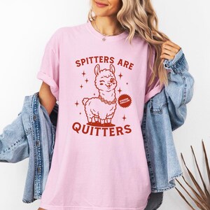 May include: Pale pink t-shirt featuring a cartoon llama with a winking eye and a necklace. The text "Spitters Are Quitters" is printed in red. The shirt is a light pink colour.