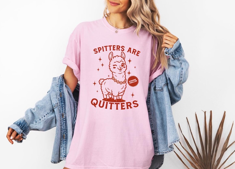May include: Pale pink t-shirt featuring a cartoon llama with a winking eye and a necklace. The text "Spitters Are Quitters" is printed in red. The shirt is a light pink colour.