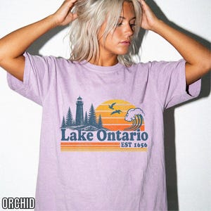 May include: An orchid-colored short-sleeve t-shirt featuring a graphic print of a lighthouse, pine trees, a large wave, and two birds against a yellow and orange sunset. The text reads Lake Ontario EST 1656 in a dark blue, retro-style font.