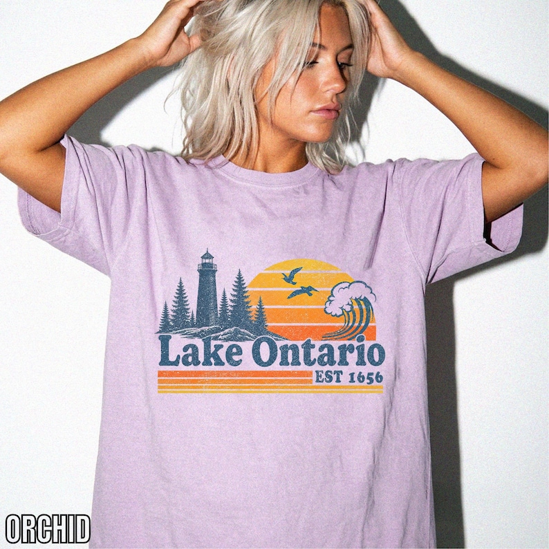 May include: An orchid-colored short-sleeve t-shirt featuring a graphic print of a lighthouse, pine trees, a large wave, and two birds against a yellow and orange sunset. The text reads Lake Ontario EST 1656 in a dark blue, retro-style font.
