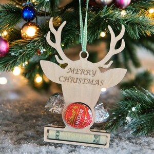 Op de afbeelding: Een houten kerstversiering in de vorm van een rendierkop met de tekst "MERRY CHRISTMAS". Het bevat een Lindor-chocolade en een klein geldbedrag. De versiering hangt aan een groen lint in een kerstboom.