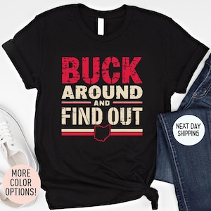 May include: Black t-shirt with the text "BUCK AROUND AND FIND OUT" in distressed red and white lettering. The Ohio state outline is featured below the text in red. The shirt is displayed with jeans and a white shoe.