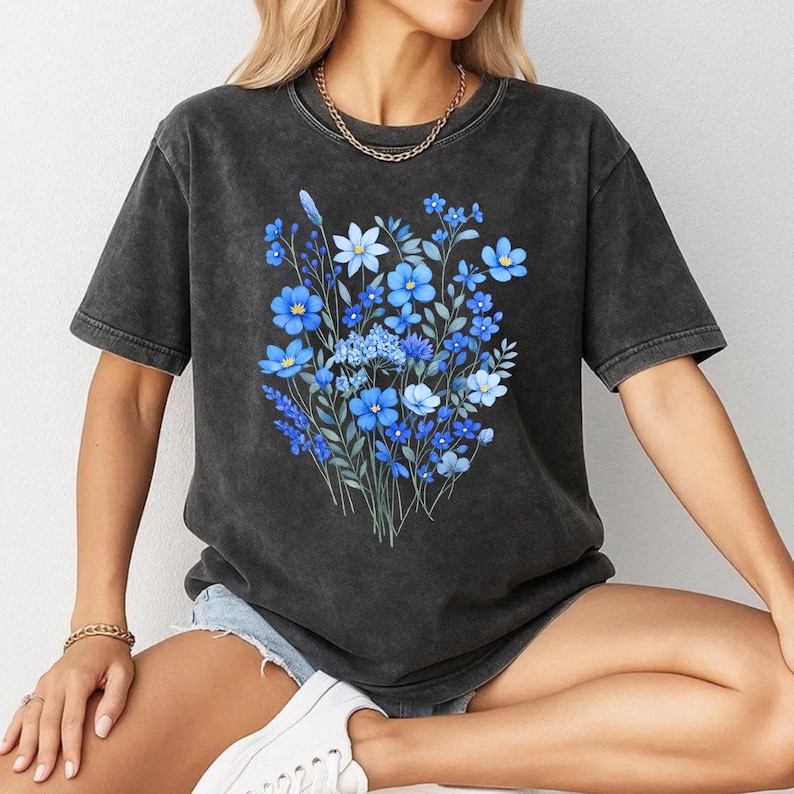 May include: Dark gray t-shirt featuring a watercolor floral design in shades of blue. The graphic includes various flower types and foliage. The shirt has a vintage, washed appearance and a classic crew neck.