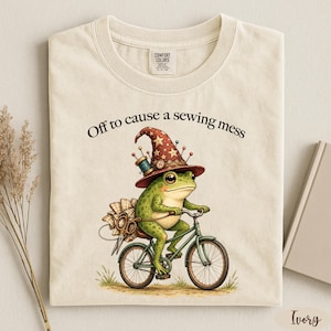 May include: An ivory t-shirt featuring a graphic of a green frog wearing a star-patterned wizard hat, riding a bicycle, and carrying sewing supplies. The text above the frog reads, Off to cause a sewing mess. The shirt has a crew neckline.