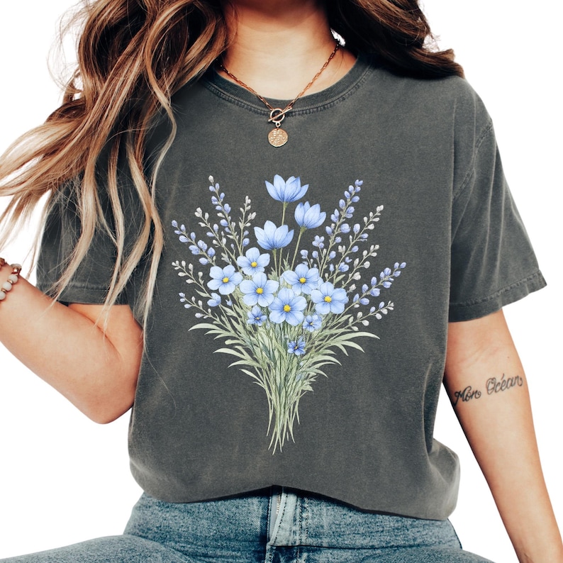 May include: Dark grey t-shirt with a floral design. The design features a bouquet of blue flowers with green stems and leaves. The t-shirt has a crew neck and short sleeves. The floral design is centered on the front.