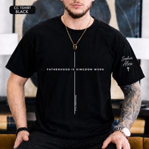 May include: A black short-sleeve t-shirt featuring a white vertical cross graphic with the text FATHERHOOD IS KINGDOM WORK and PROVERBS 22:6. The left sleeve displays the names Sophia Alex above a small cross symbol in white lettering.