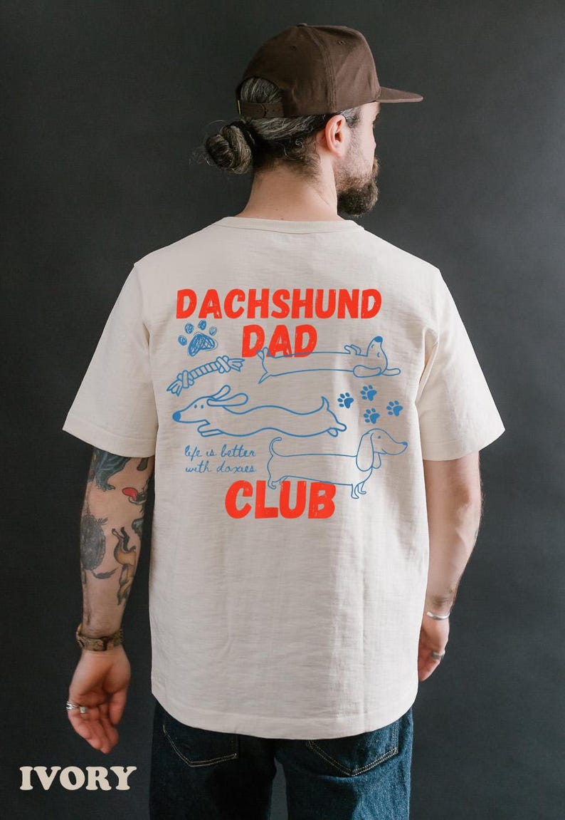 May include: Ivory t-shirt with the words "DACHSHUND DAD CLUB" in red and blue. The shirt features illustrations of dachshunds and paw prints. The text "life is better with doxies" is also printed on the shirt.