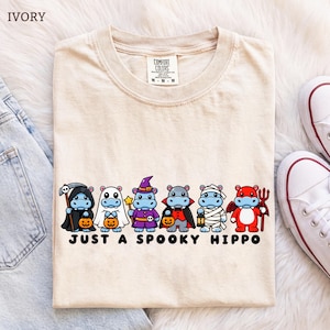Op de afbeelding: Een ivoorkleurig t-shirt met een afbeelding van zes cartoon-nijlpaarden in verschillende Halloween-kostuums, waaronder een Magere Hein, geest, heks, vampier, mummie en duivel. De tekst onder de nijlpaarden luidt "JUST A SPOOKY HIPPO."